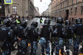 Russiske sikkerhetsstyrker st&aring;r oppstilt foran demonstranter og mennesker i en gate. 