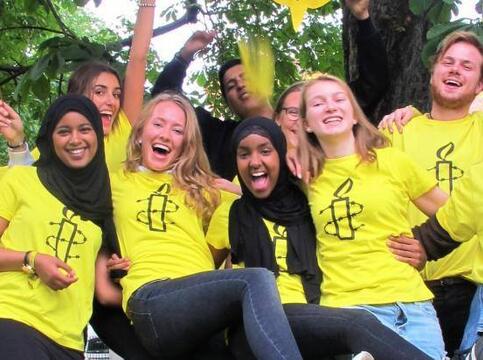 En gruppe jenter og gutter i gule Amnesty T-skjorter med Amnestys logo p&aring;, jubler og rekker armene i v&aelig;ret. De har gule Amnesty-flagg som de vifter med. Noen av jentene har langt lyst h&aring;r, guttene har kort lyst eller m&oslash;rkt h&aring;r og to av jentene har svart hijab.